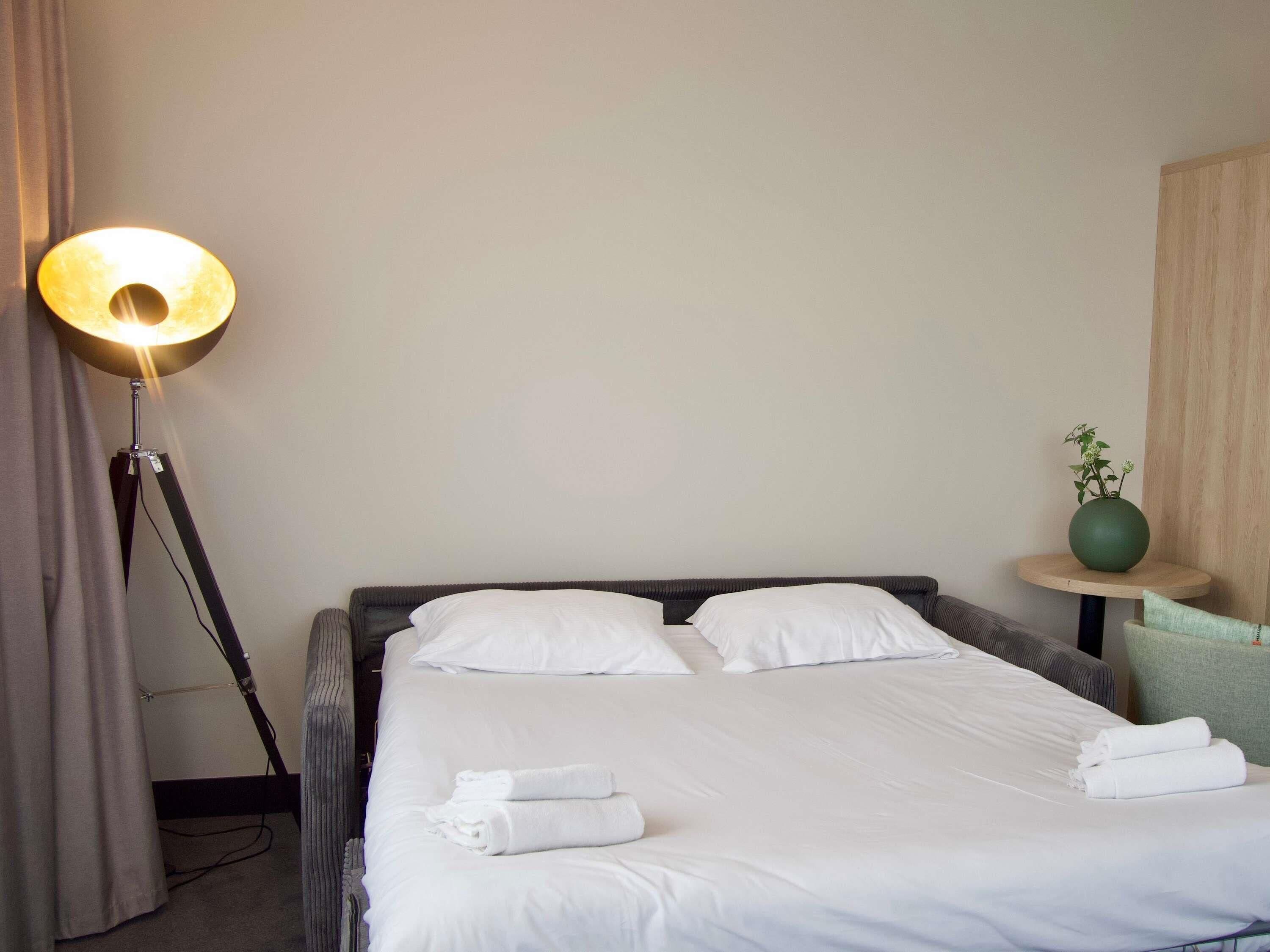 A neatly made bed with white linens and towels, a floor lamp, a small round table with a green vase, and a wooden wall panel.