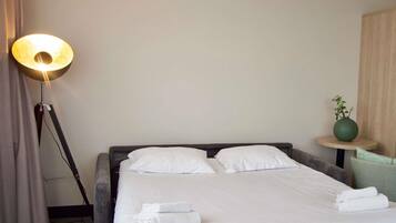 A neatly made bed with white linens and towels, a floor lamp, a small round table with a green vase, and a wooden wall panel.