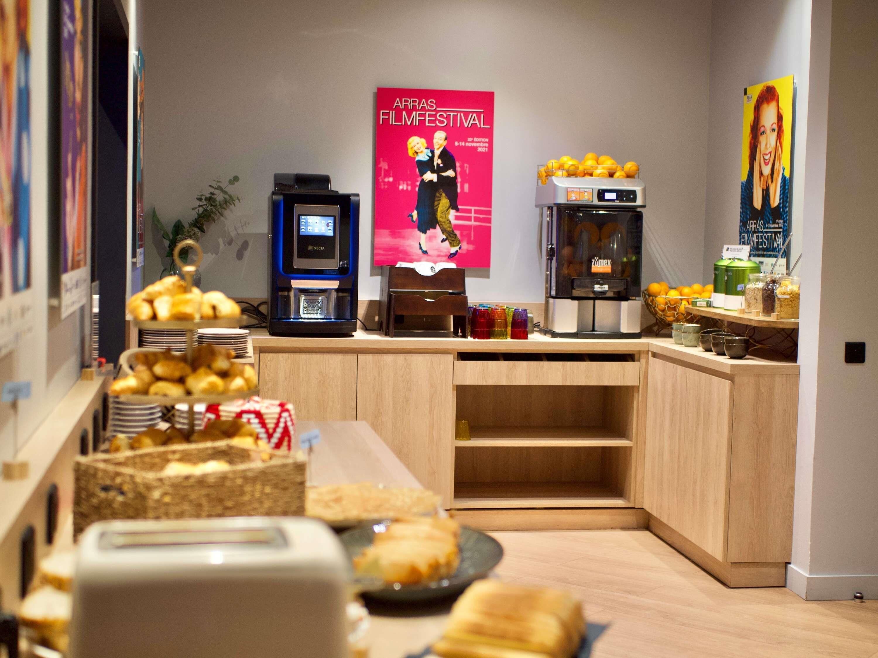 A kitchenette with a coffee machine, a fruit dispenser, and a poster for the Arras Film Festival.