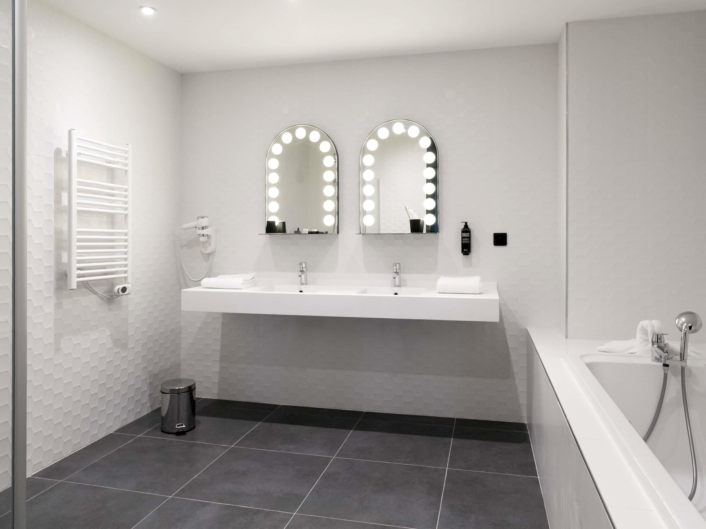 Een moderne badkamer met twee wastafels, een spiegel met verlichting, een handdoekhouder, een prullenbak en een bad.