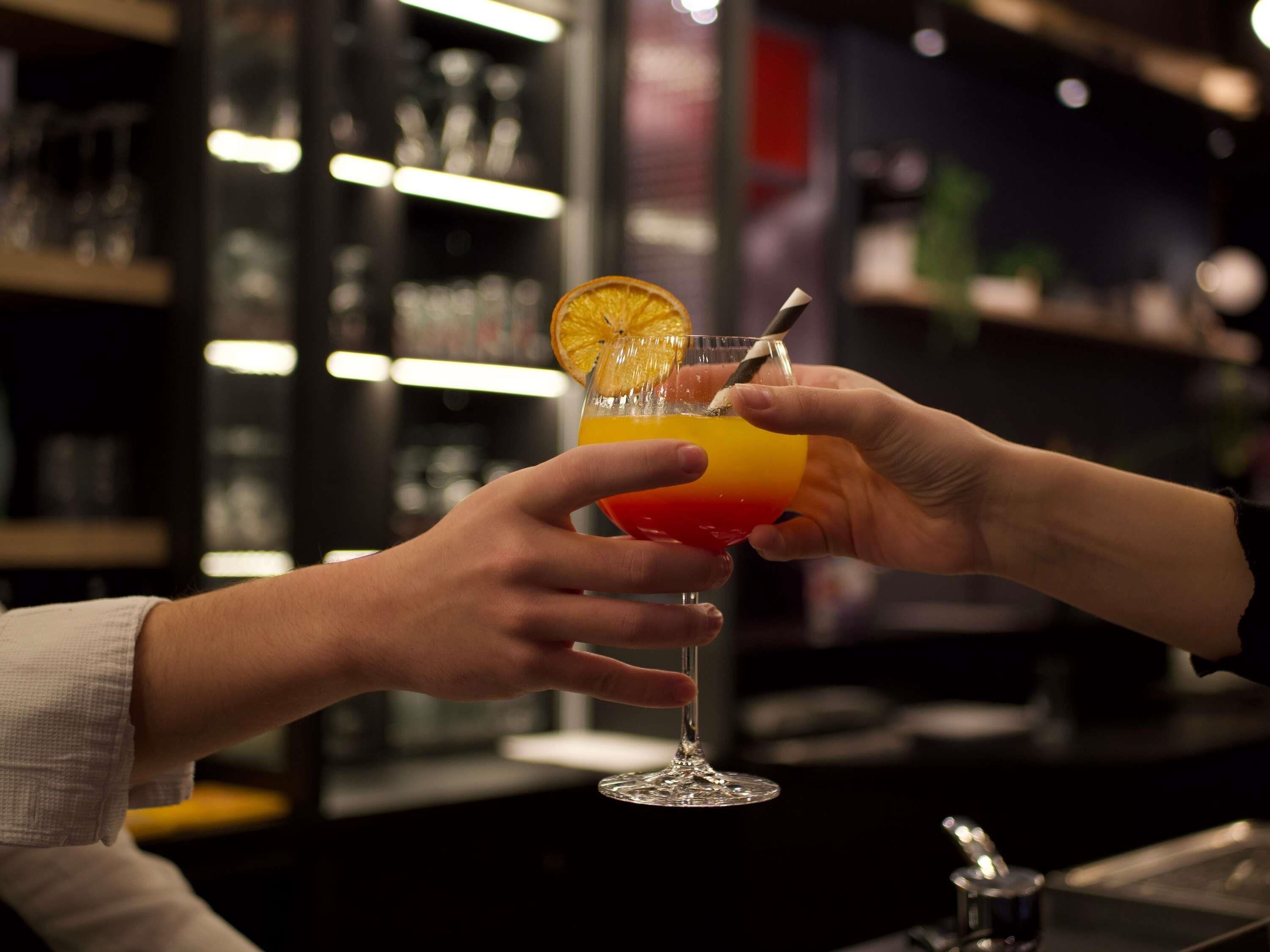 Two hands holding a cocktail glass with a gradient color and an orange slice garnish.