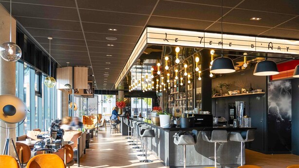 Un restaurante moderno con zona de bar, techo alto con iluminación colgante y vista a la calle.