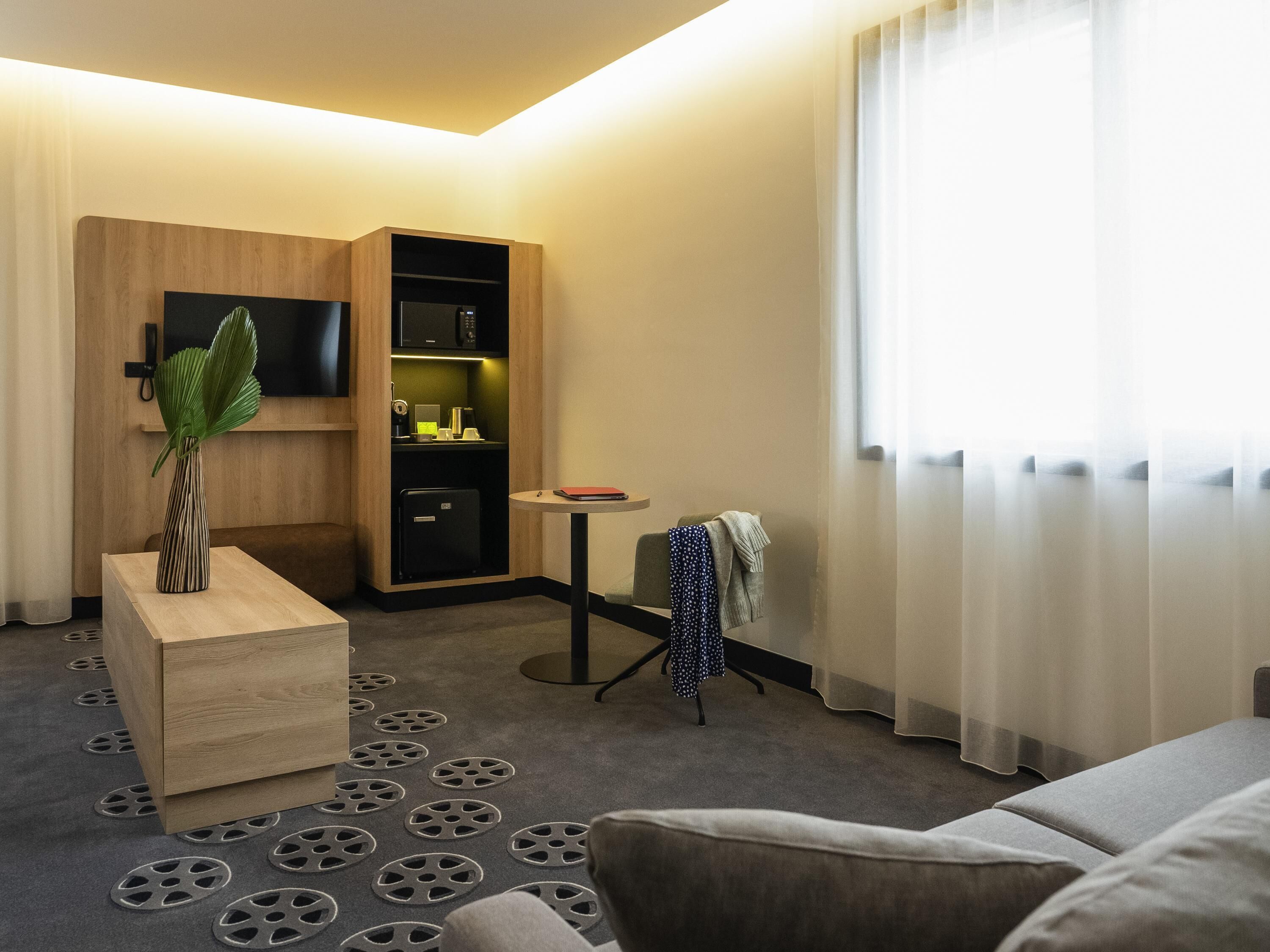 A hotel room with a sofa, a small table, a TV, a minibar, and a window with curtains.