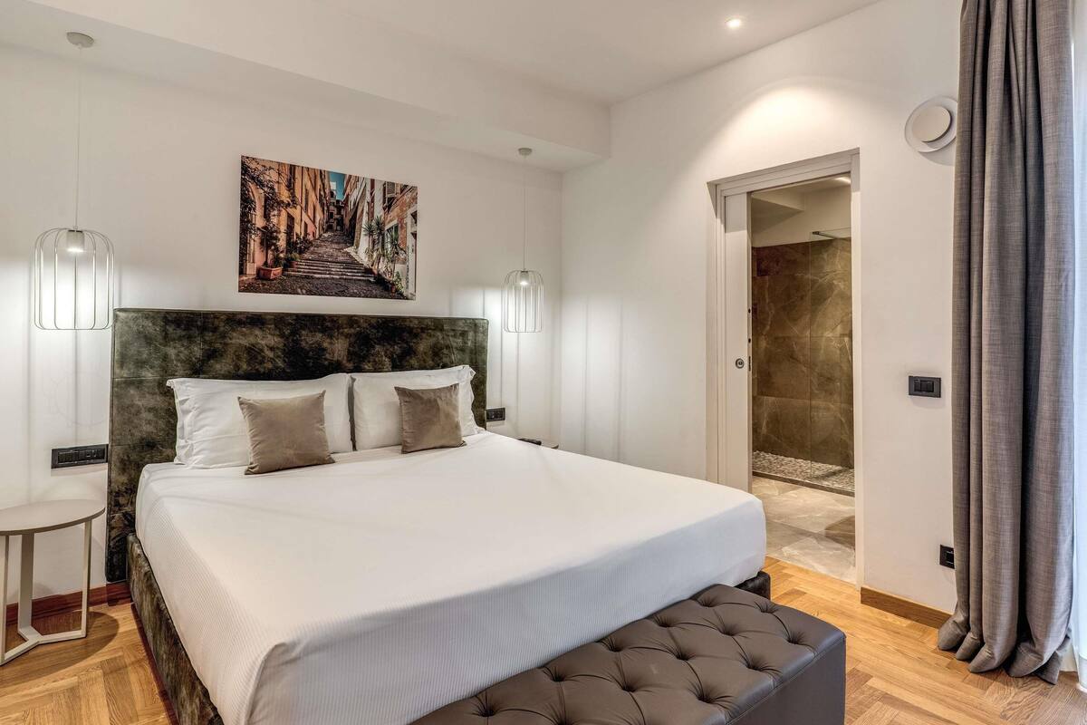 Ein modernes Schlafzimmer mit einem großen Bett, einem Kopfteil mit strukturierter Optik, einem Wandbild und einem sichtbaren Duschbereich durch eine geöffnete Tür.