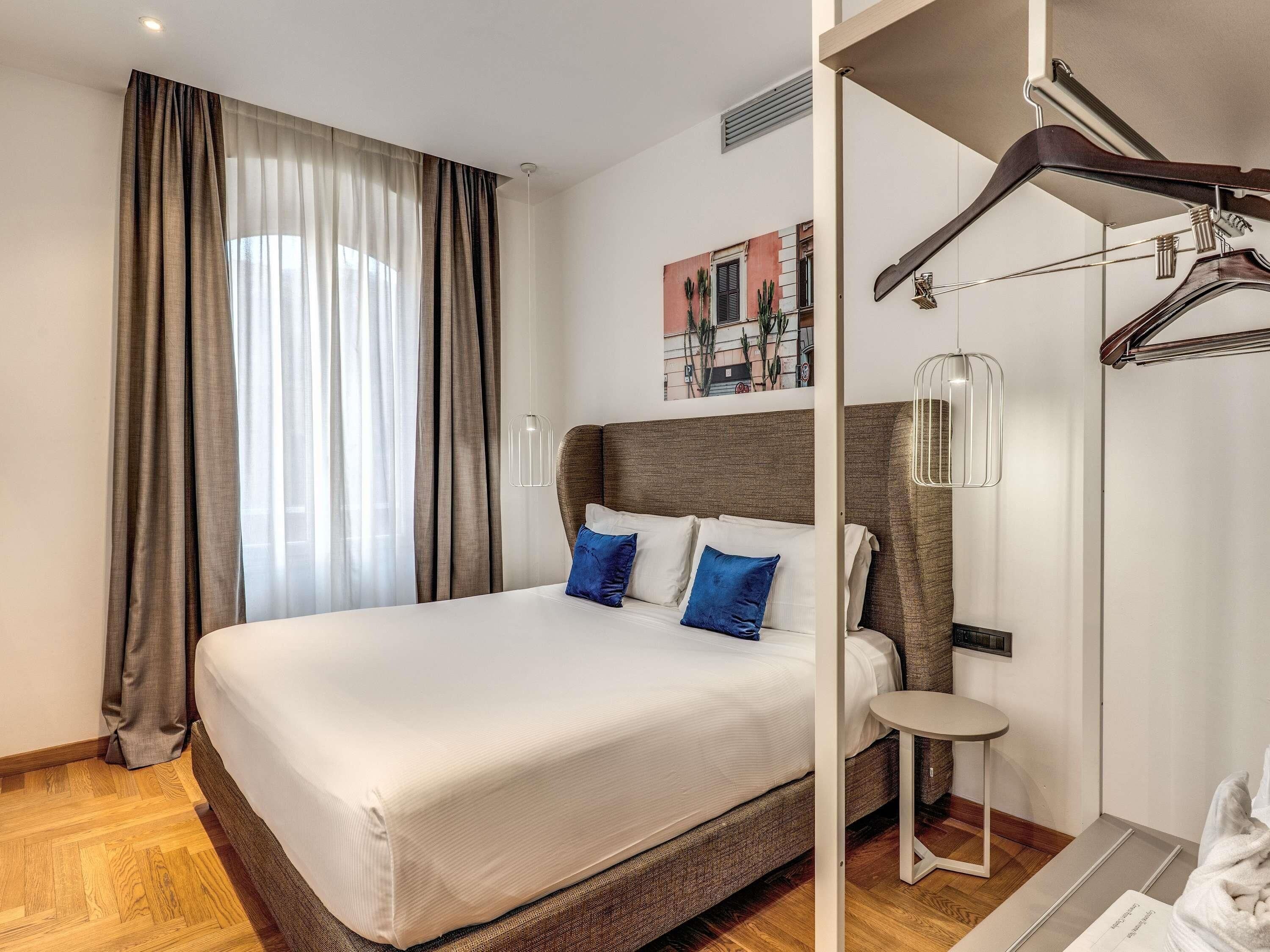 Habitación de hotel con una cama grande, una mesita pequeña, un perchero y un cuadro en la pared.