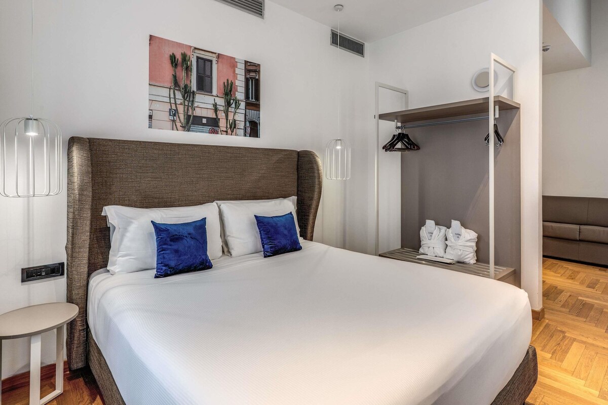 Quarto de hotel moderno com uma cama grande, mesa de cabeceira, guarda-roupa e um quadro na parede.