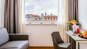 Hotelkamer met uitzicht op de stad, een bank, een stoel, een tafel met een fruitmand en een bloevas.
