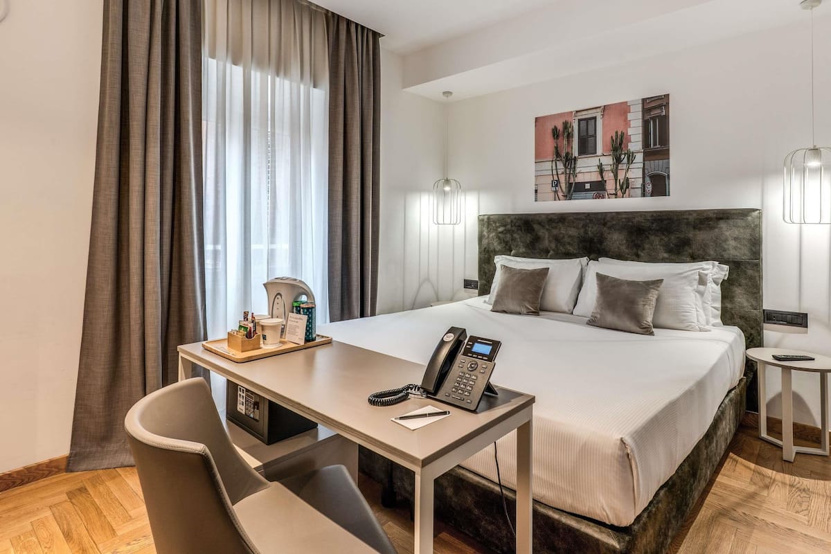 Ein Hotelzimmer mit Bett, Schreibtisch mit Telefon, Stuhl, kleinem Tisch mit Tasse und Tablett sowie einer Wandlampe.