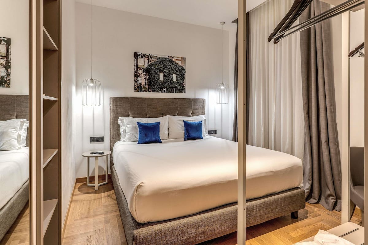 Ein Hotelzimmer mit Bett, Nachttischen, einer Pendelleuchte, einem Wandbild und einem Kleiderschrank mit Spiegel.