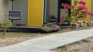 A building with two yellow sliding doors, a small patio with a bench, a purple umbrella, and a pink flowering plant. - Cosy studio overlooking the rock, 5 minutes from the beach on foot (Le Diamant)