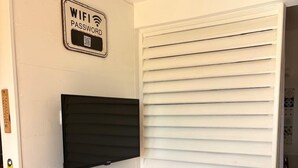 A small dining area with a table and chairs, a TV mounted on the wall, and a sign indicating a WiFi password. - Cosy studio overlooking the rock, 5 minutes from the beach on foot (Le Diamant)