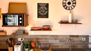A kitchen with a microwave, toaster, and sink. There are shelves with dishes and a clock on the wall. - Cosy studio overlooking the rock, 5 minutes from the beach on foot (Le Diamant)