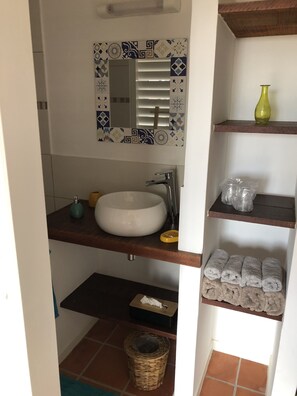 A bathroom with a white sink, a mirror with a decorative frame, and shelves with towels and toiletries. - Cozy studio with a view of the cliffs, a 5-minute walk to the beach (Le Diamant)