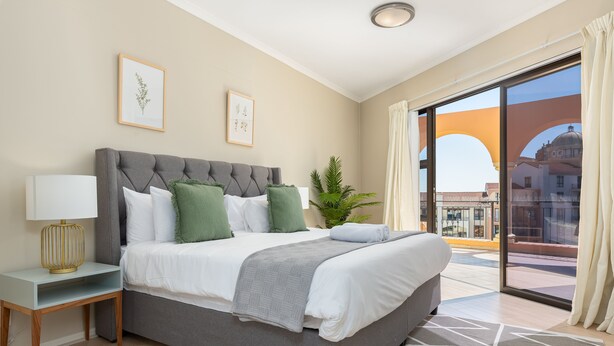 A modern bedroom with a large bed, a nightstand, a lamp, and a view of the city through a balcony. - Island Club 410 (Cape Town)
