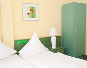 Ein Hotelzimmer mit Bett, Kissen, einem grünen Nachttisch mit Lampe, einem grünen Kleiderschrank und einem gerahmten Bild an der Wand.