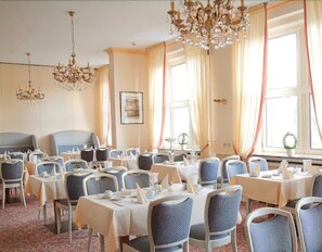 Ein Hotelrestaurant mit mehreren Tischen, Stühlen, Kronleuchtern und großen Fenstern mit Vorhängen.