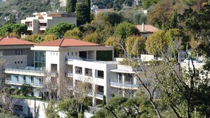 A modern multi-story building with balconies, surrounded by trees and hills in the background.