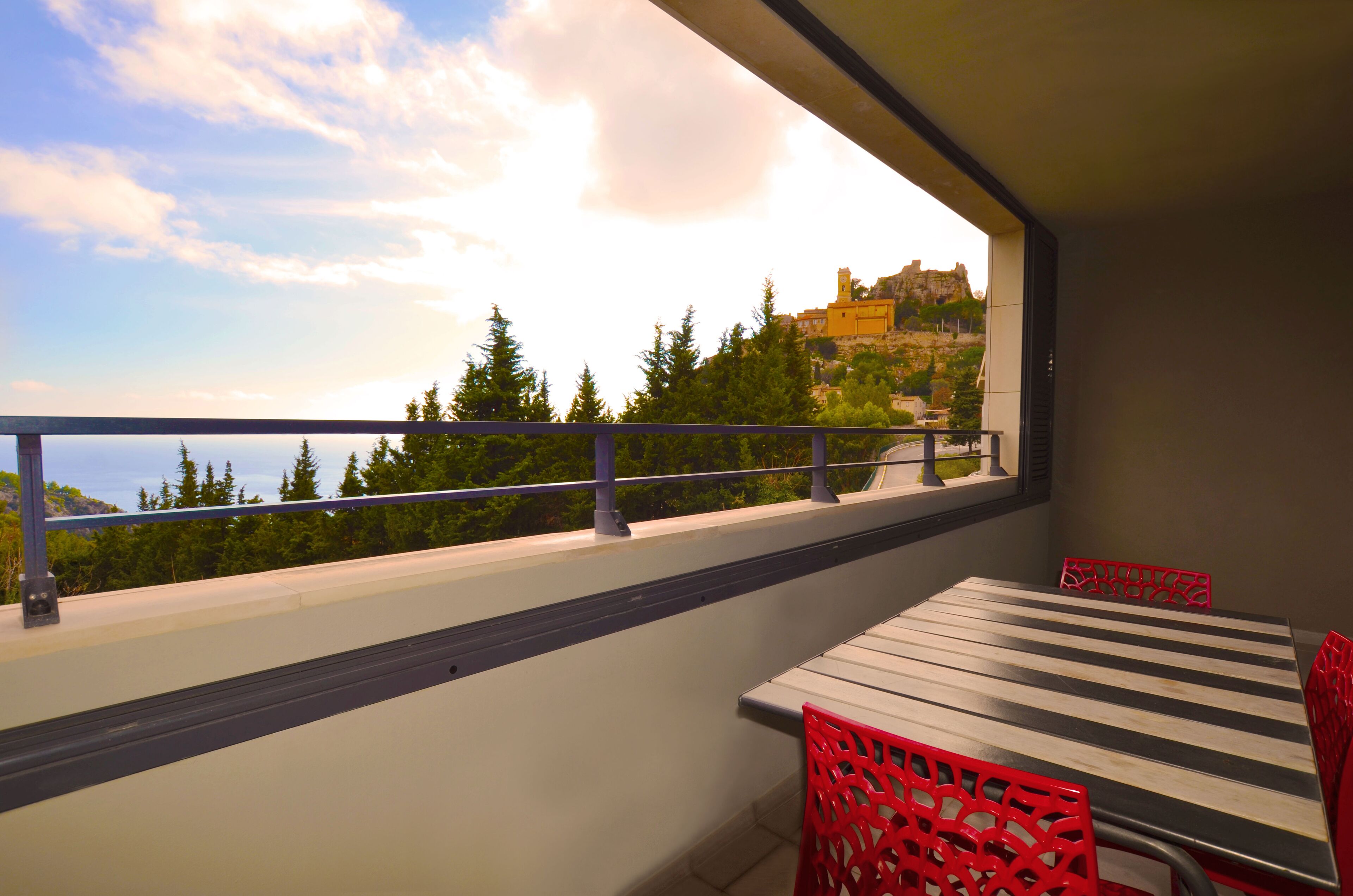 A balcony with a table and red chairs, overlooking a mountainous landscape with trees and a building.