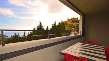 A balcony with a table and red chairs, overlooking a mountainous landscape with trees and a building.