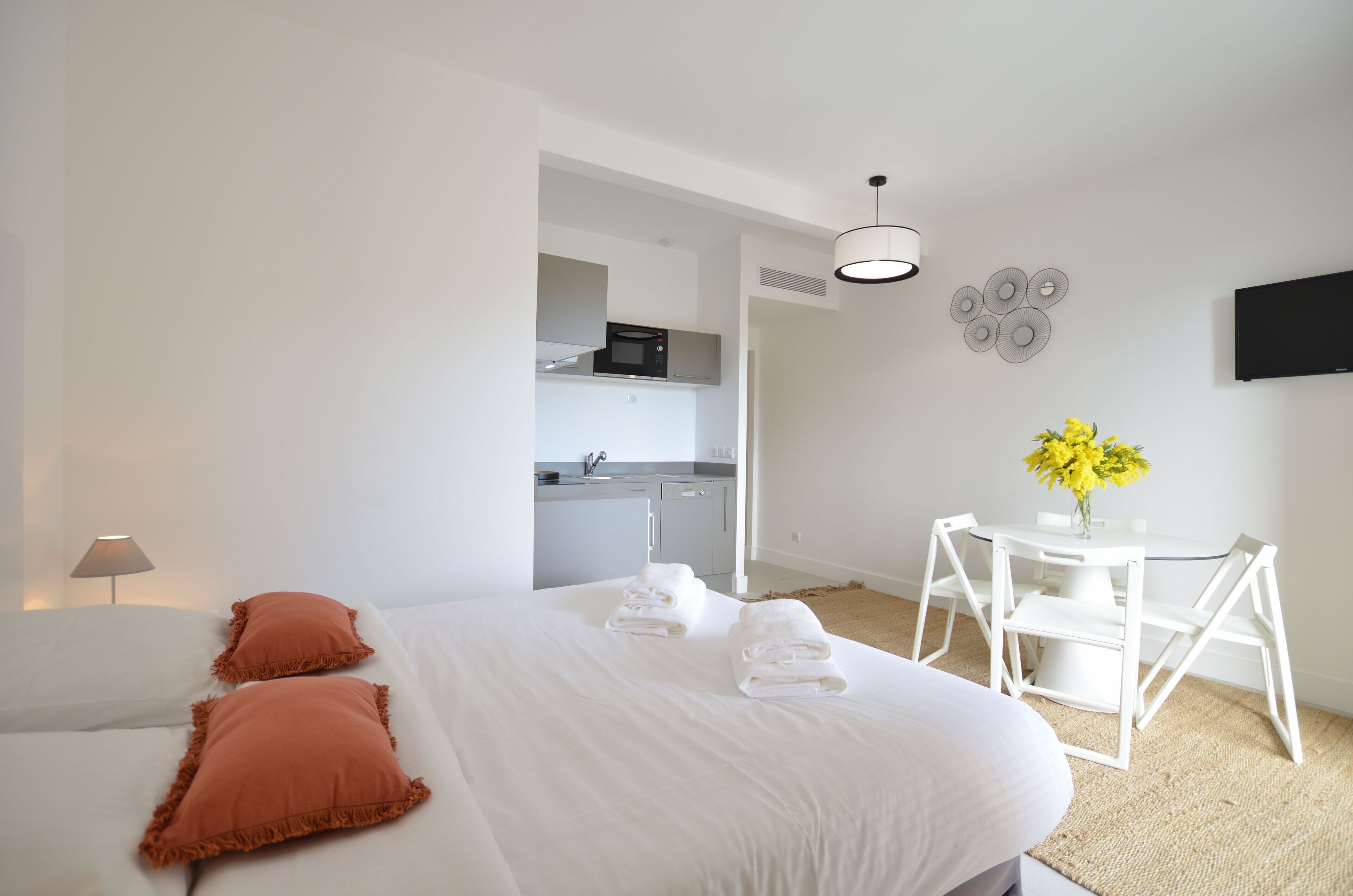 A modern hotel room with a neatly made bed, a small dining table with chairs, a kitchen area, and a flat-screen TV mounted on the wall.