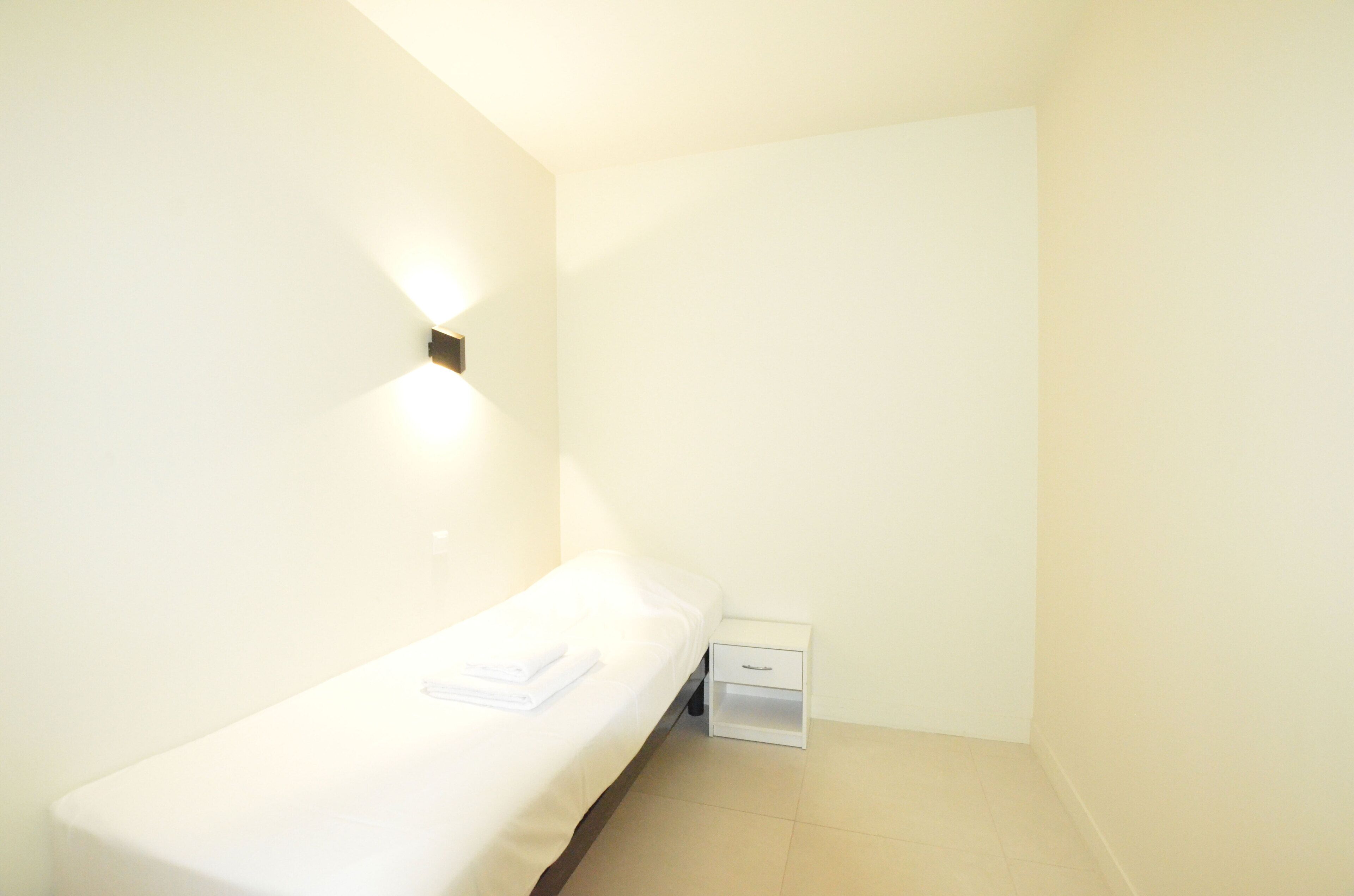 A small, single-bed room with a white bed, a nightstand with a drawer, and a wall-mounted light fixture.