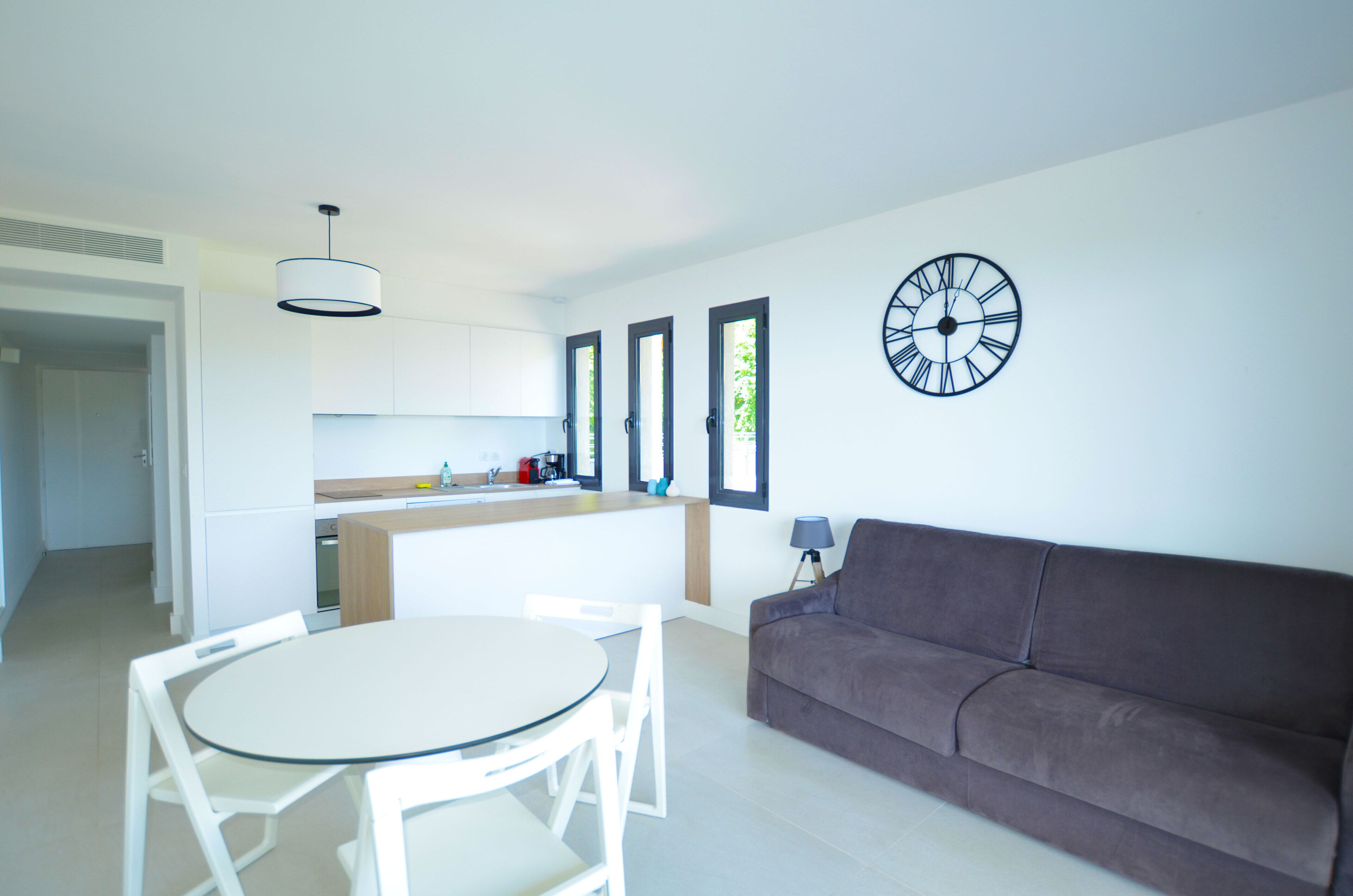 Una cocina moderna con armarios blancos, una mesa de comedor con sillas, un sofá marrón y un reloj de pared.