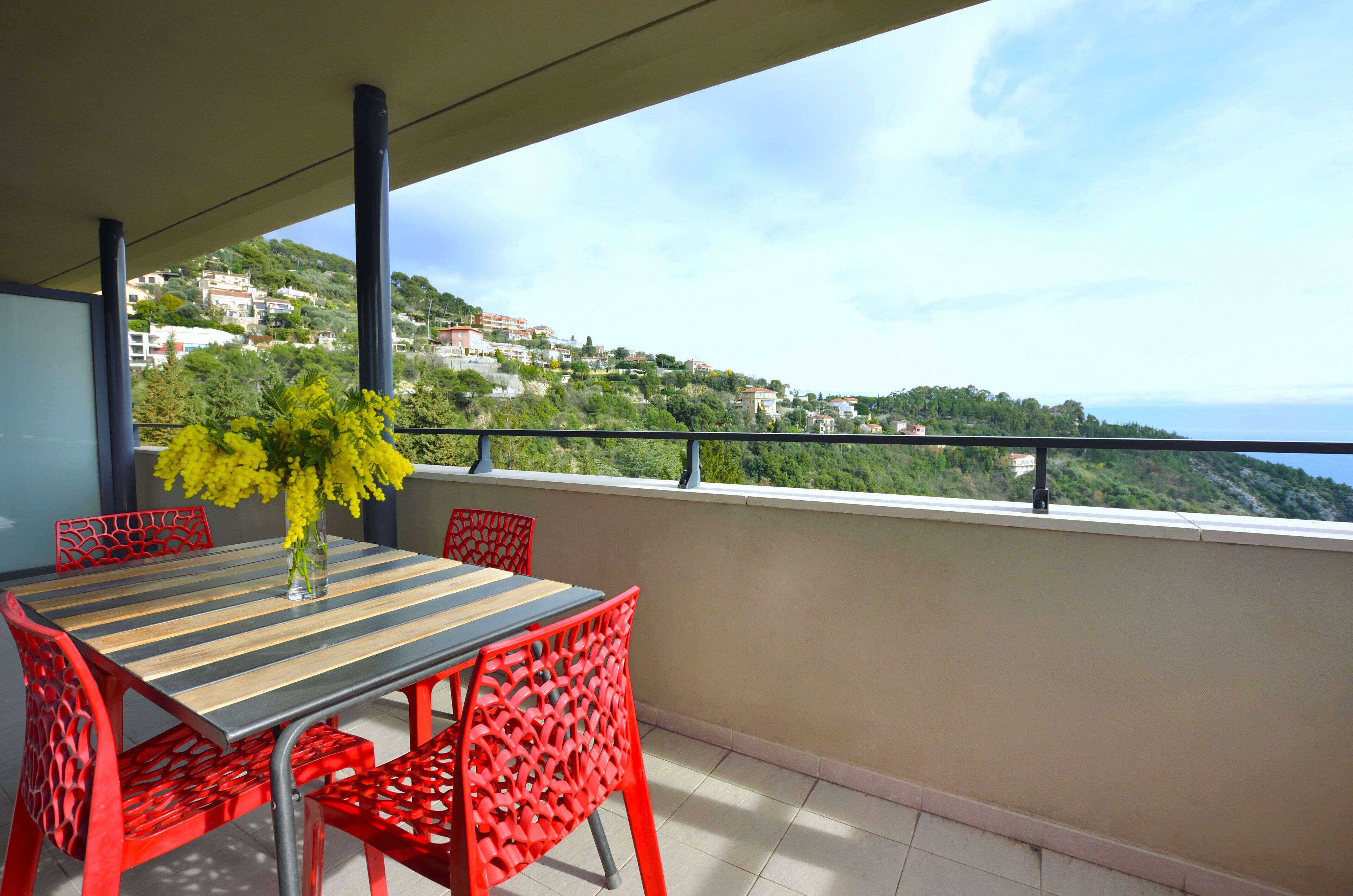 A balcony with a striped table and red chairs, overlooking a hillside with buildings.