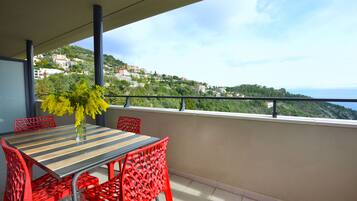 A balcony with a striped table and red chairs, overlooking a hillside with buildings.
