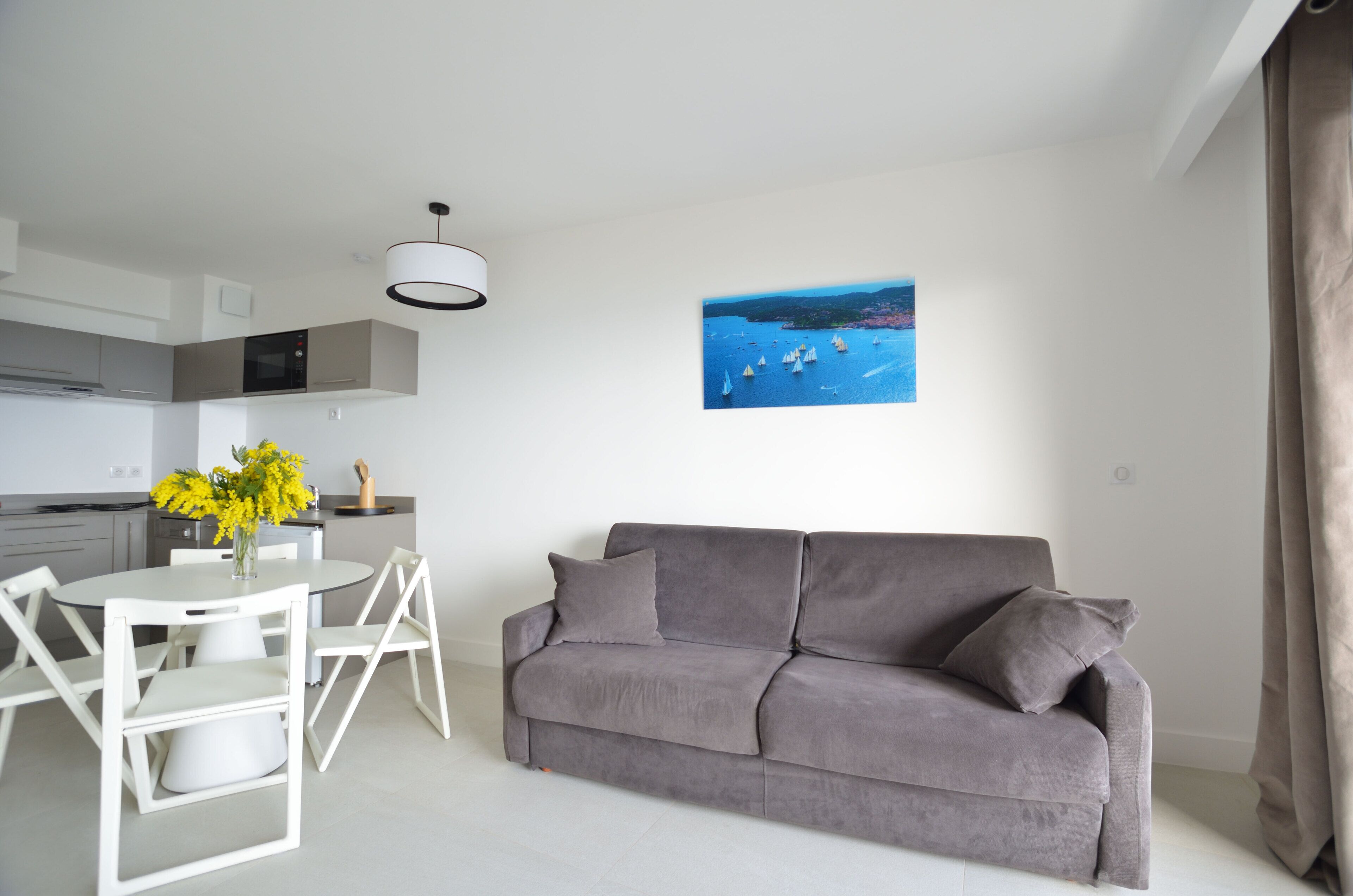 A modern living room with a grey sofa, a white dining table with chairs, and a painting of a coastal scene.