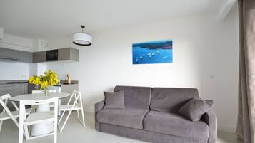 A modern living room with a grey sofa, a white dining table with chairs, and a painting of a coastal scene.