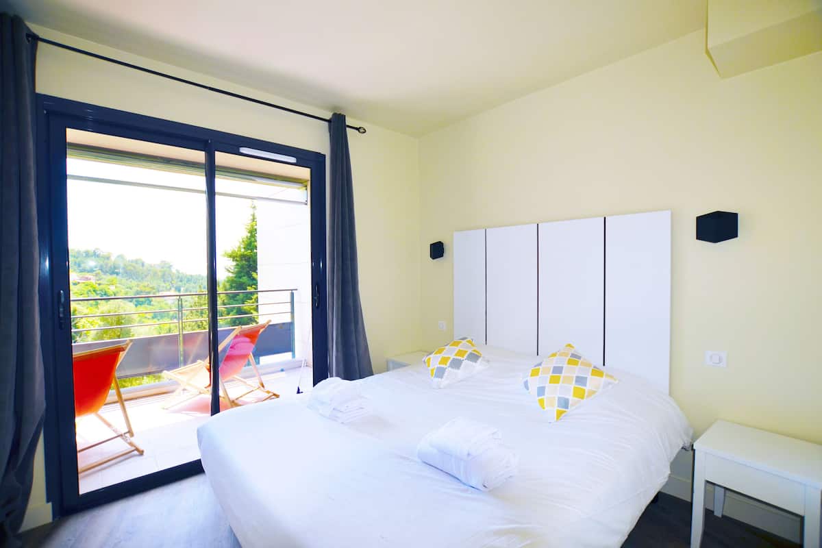 Un dormitorio con una cama grande, dos almohadas, una mesita de noche y vistas a un balcón con sillas.