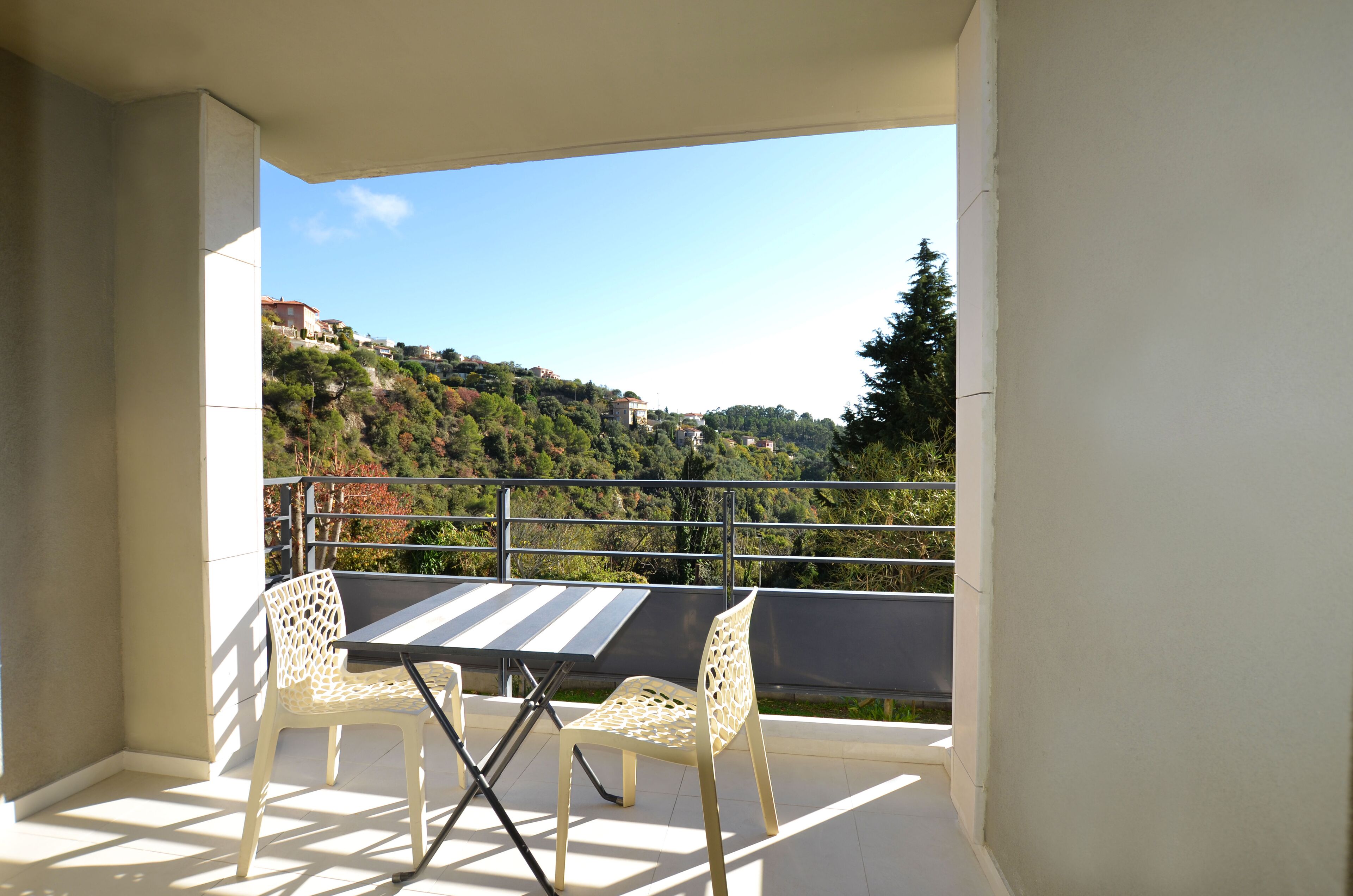 A balcony with a table and two chairs, overlooking a hillside with buildings and trees.