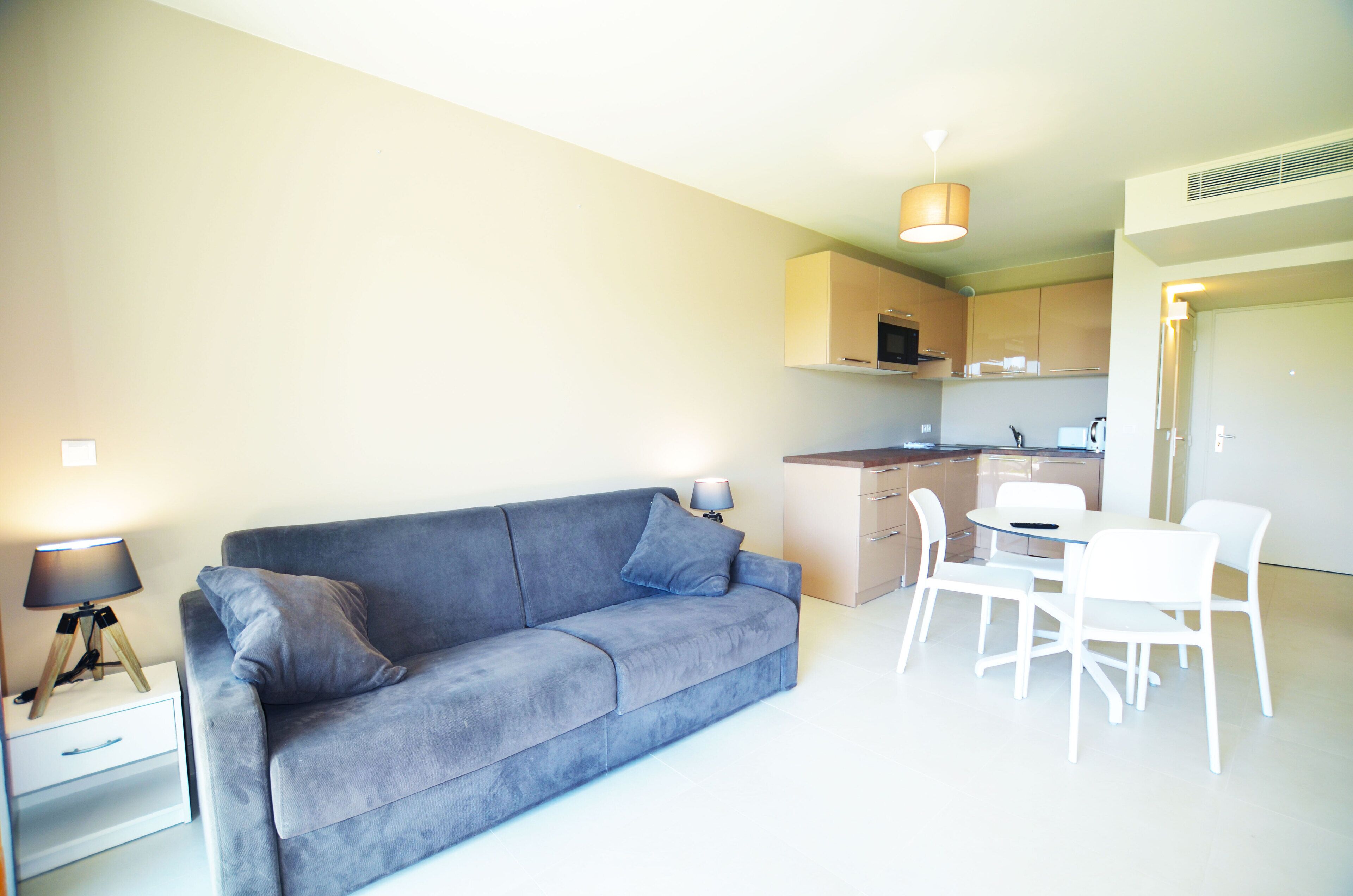 Un espace de vie moderne avec un canapé gris, une petite table entourée de chaises, une lampe et, au fond, une kitchenette.