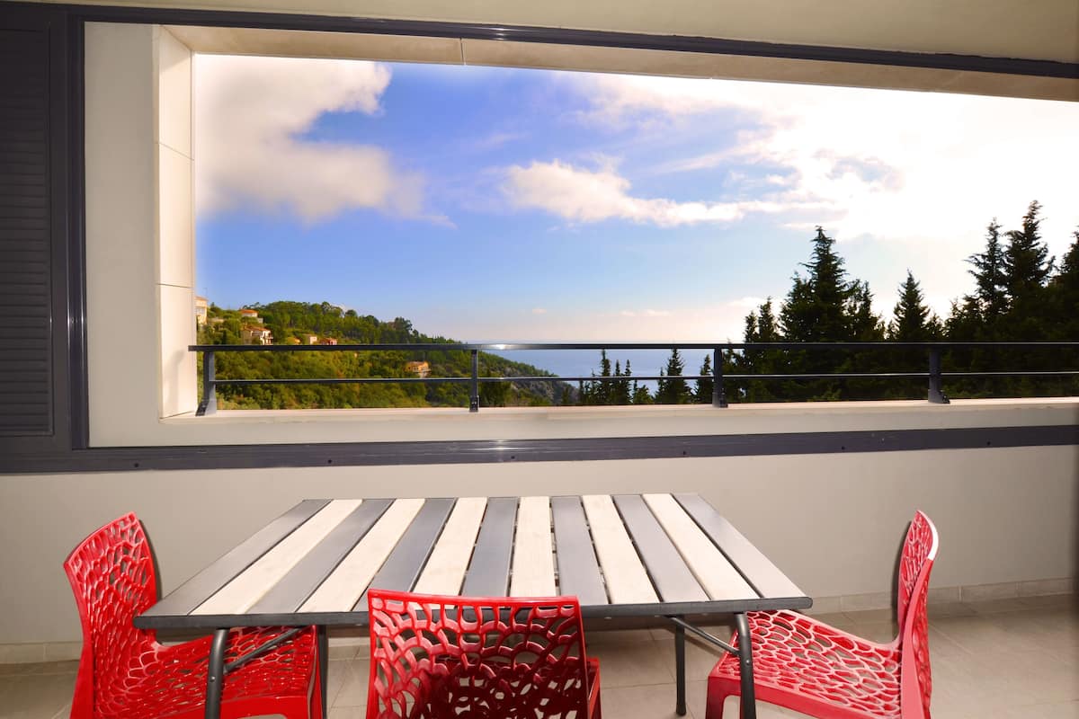 Un balcón con mesa y sillas, con vista a un paisaje escénico de árboles y una costa lejana.