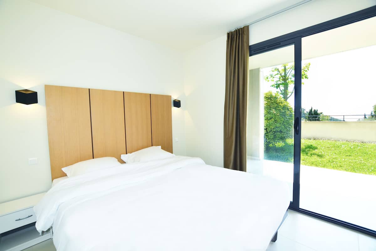 Un dormitorio moderno con una cama grande, cabecera de madera y puerta corrediza de vidrio que da a un balcón con vistas a la vegetación.