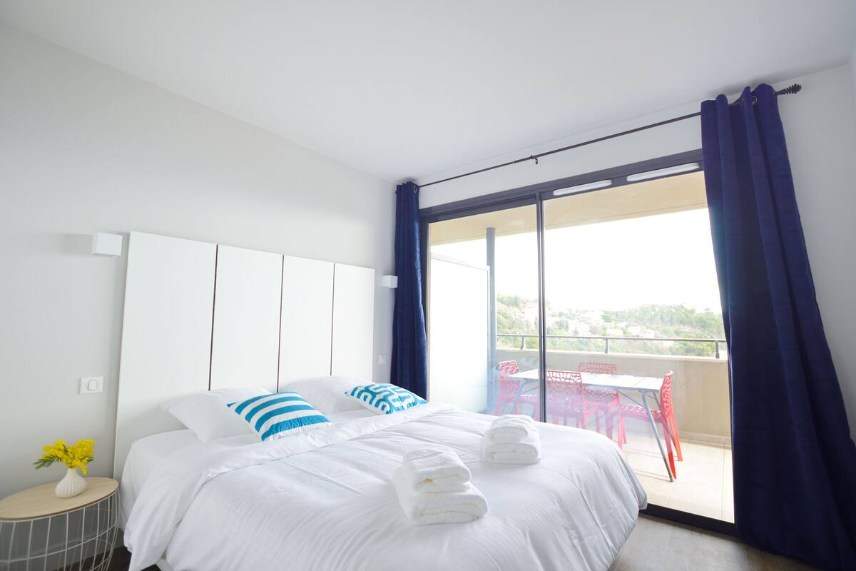 Habitación de hotel con cama, una mesita con un florero y un balcón con sillas rojas.