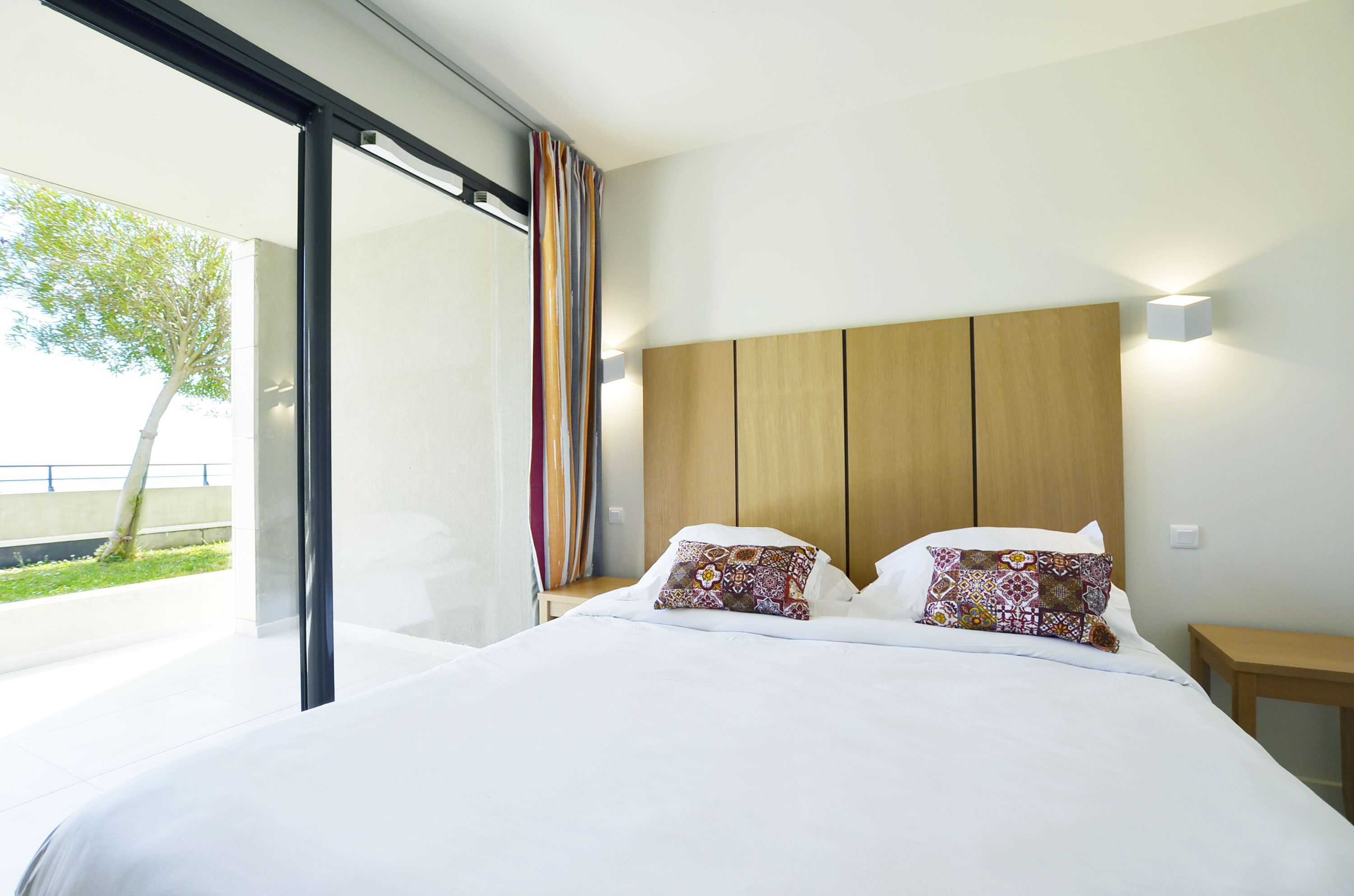 A modern hotel room with a large bed, a wooden headboard, and a view of greenery outside.