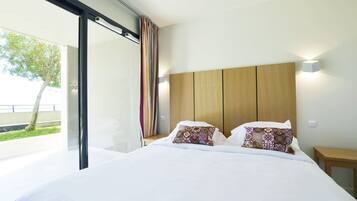 A modern hotel room with a large bed, a wooden headboard, and a view of greenery outside.