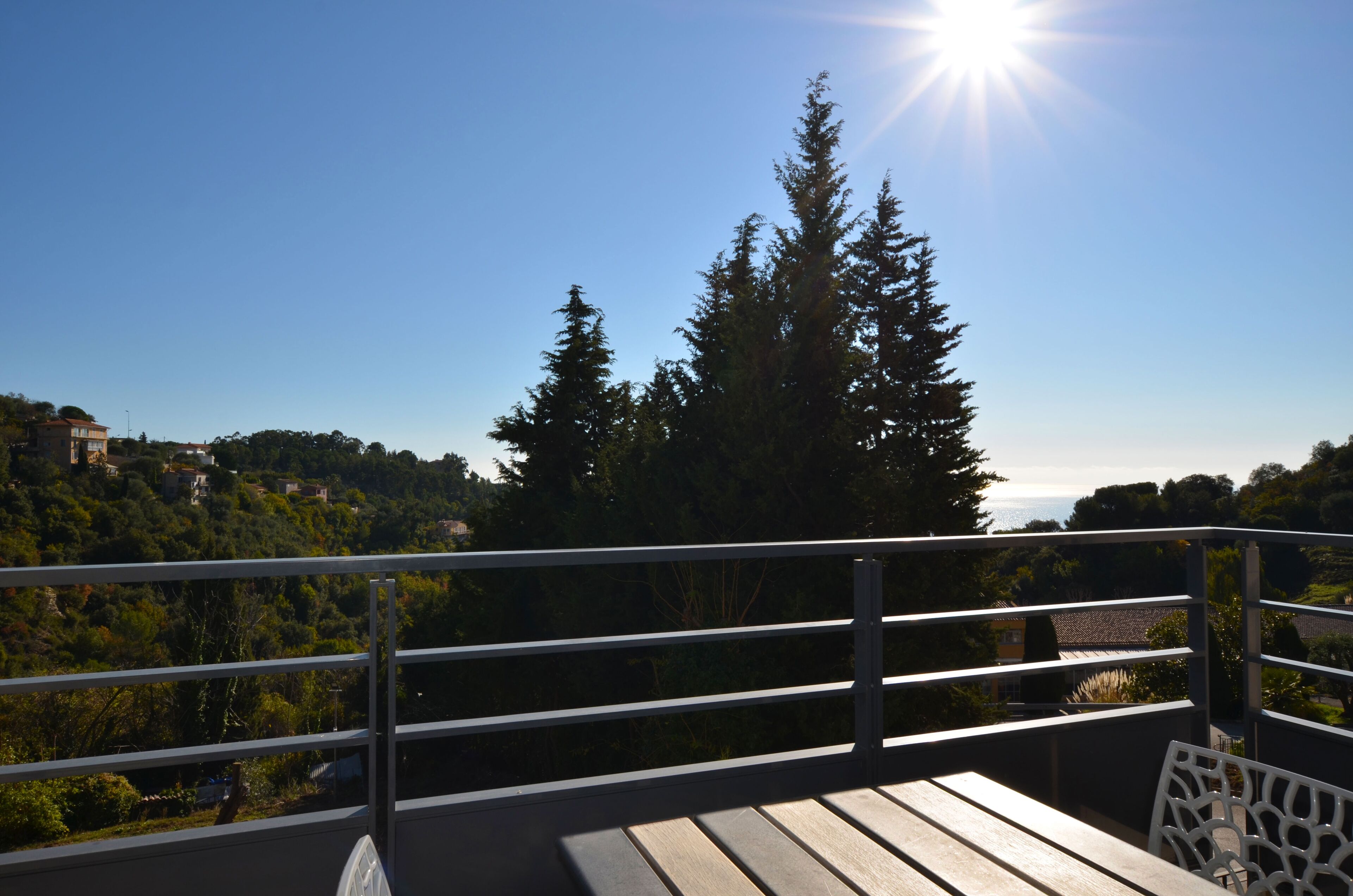 A balcony with a metal railing overlooking a forested area and a distant view of a town.