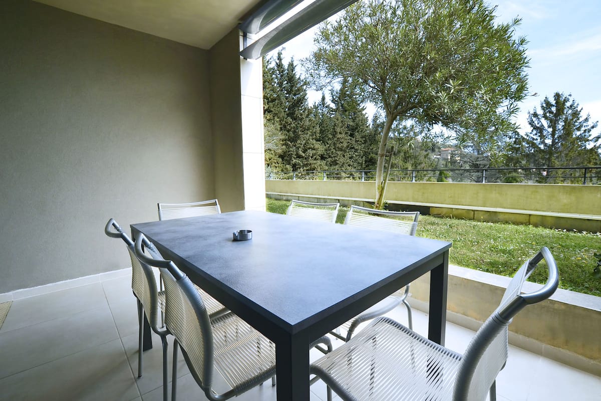 Un balcón con mesa y sillas, vistas a árboles y una valla, y cielo despejado.