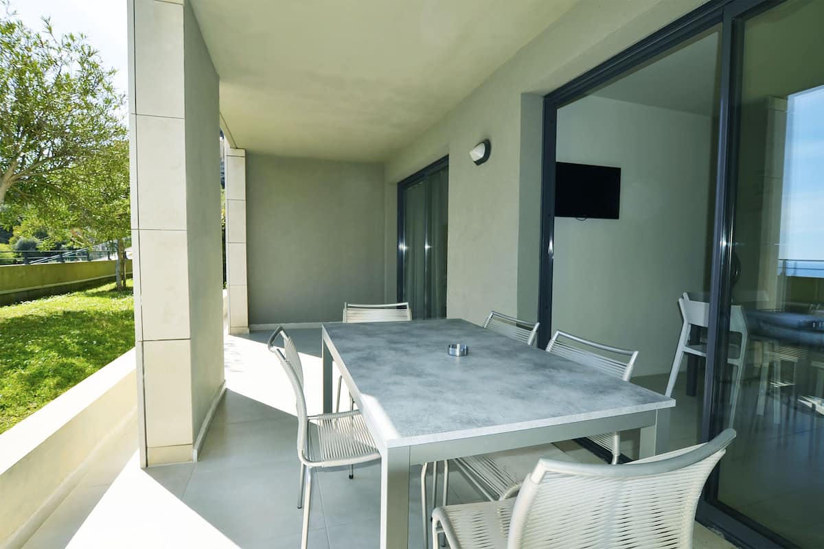 Un área de comedor exterior moderna con una mesa rectangular y sillas blancas, una puerta de cristal que da a un balcón y un televisor de pantalla plana montado en la pared.