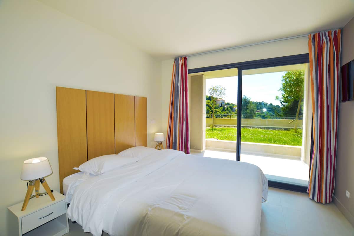 Un dormitorio con una cama grande, un cabecero de madera, una mesita de noche con una lámpara y vistas a un jardín a través de una puerta corrediza de vidrio.