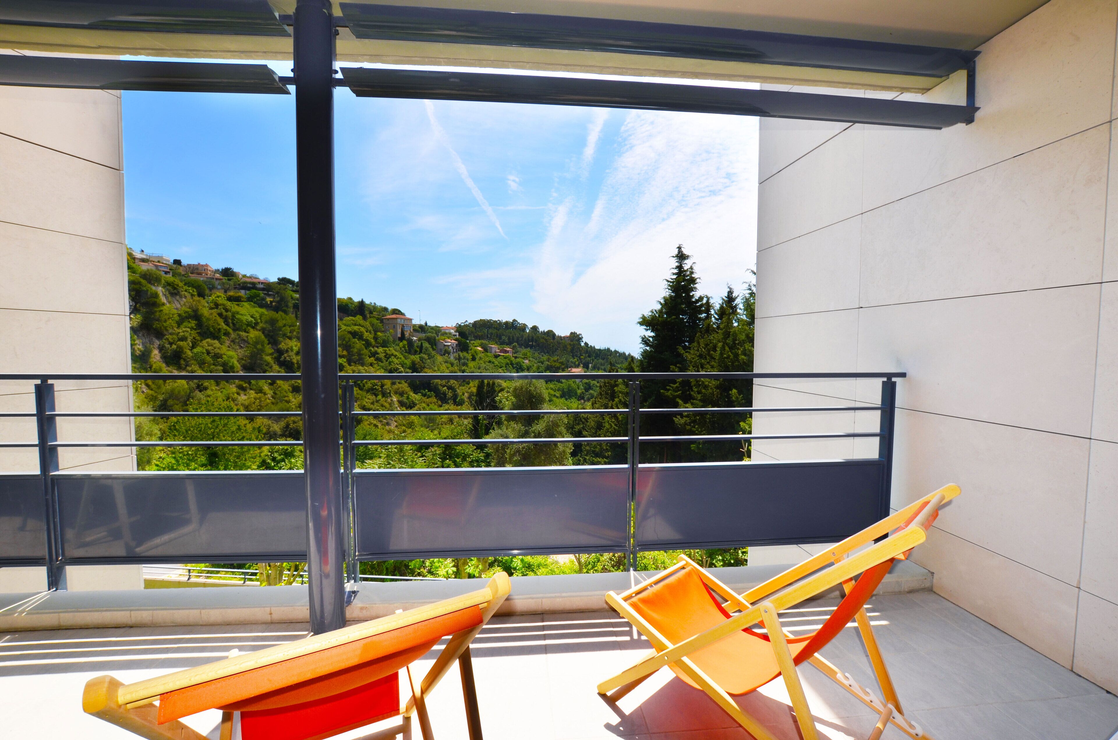 A balcony with two wooden lounge chairs, a metal railing, and a view of a green hillside.