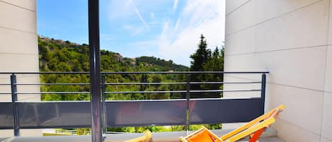 Un balcón con dos sillones de madera, barandilla metálica y vista a una ladera verde.