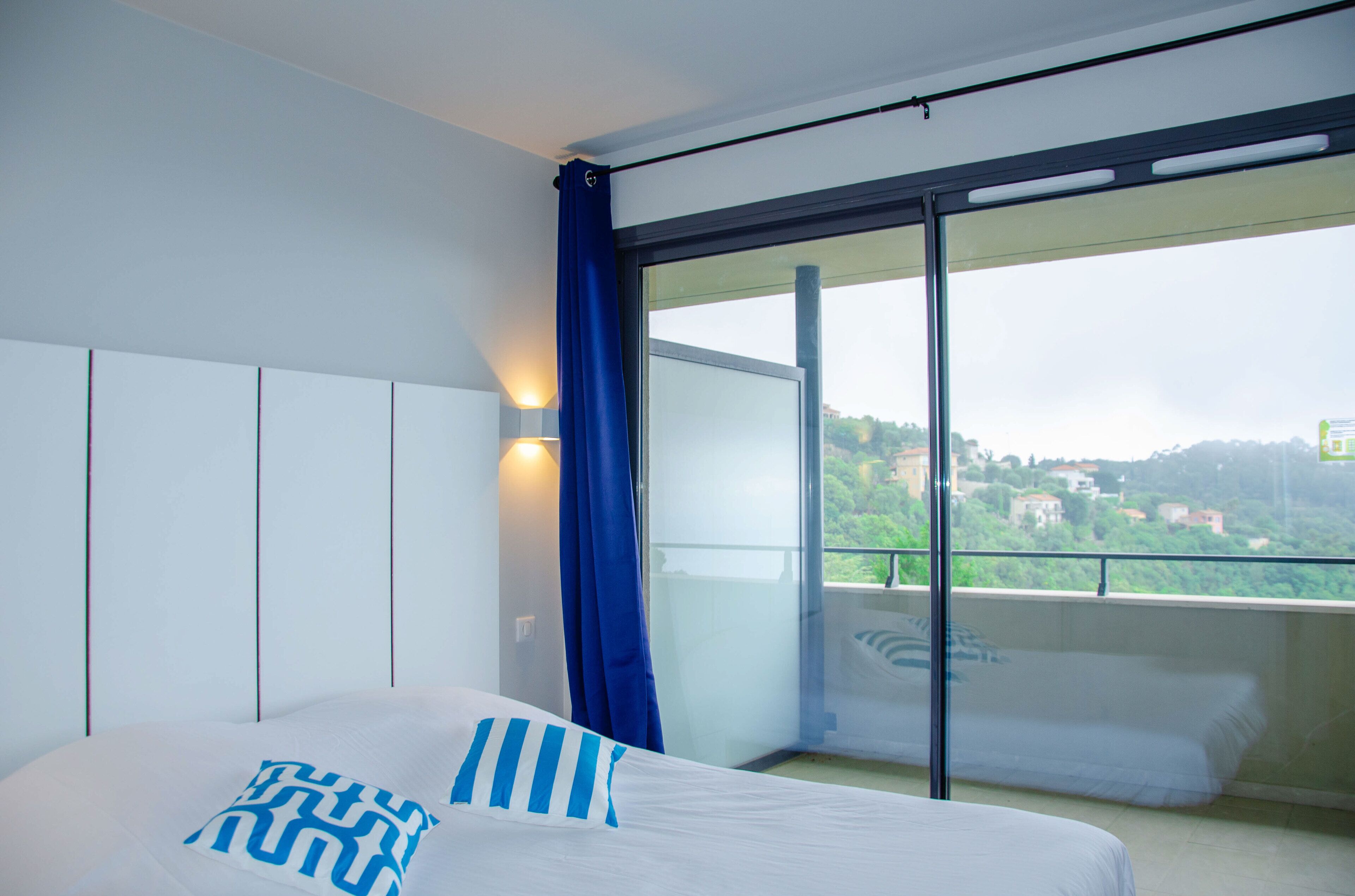 A bedroom with a bed, a large window, and a view of a balcony.