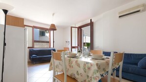 Ein Essbereich mit einem Tisch, gedeckt für vier Personen, umgeben von Stühlen. Im Hintergrund befindet sich eine Couch und eine Klimaanlage an der Wand.