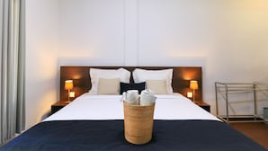 A hotel room with a bed, two bedside lamps, a bedside table, a nightstand, a wall-mounted shelf, and a wicker basket with towels. - Kak Garden Inn by Zuzu (Kuta)