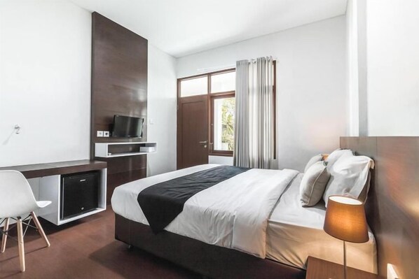 A modern hotel room with a bed, a desk, a television, and a window with curtains. - Kak Garden Inn by Zuzu (Kuta)