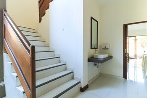 A staircase with a wooden handrail leading to a landing, a white wall with a mirror and a sink, and a door leading to an outdoor area. - Kak Garden Inn by Zuzu (Kuta)