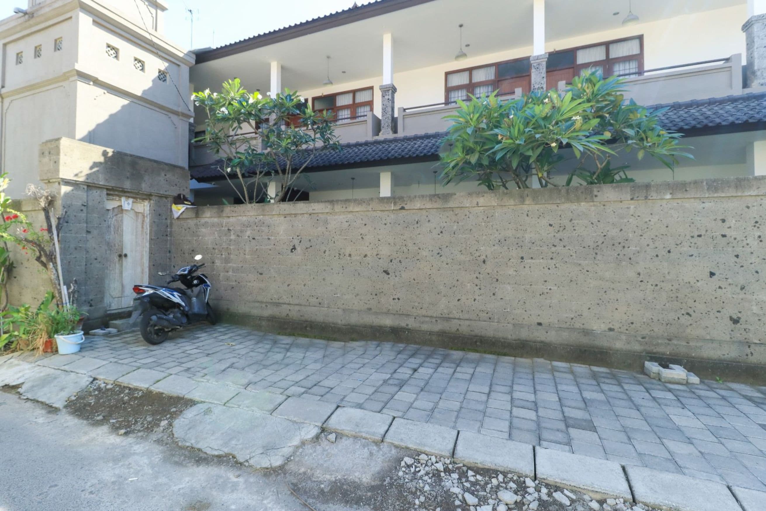 Un muro di cemento stretto con un vialetto lastricato, uno scooter parcheggiato e un edificio con finestre e un tetto piastrellato sullo sfondo.